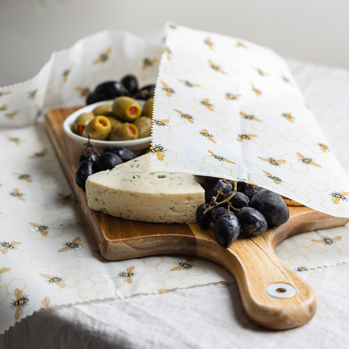 White Beeswax Wrap with tiny bees patterned, is wrapping a pre-made charcuterie board on a wooden serving tray. Including grapes, cheese and olives. The wrap is covering half of the platter and the idea is that you can make and store it ahead of time.