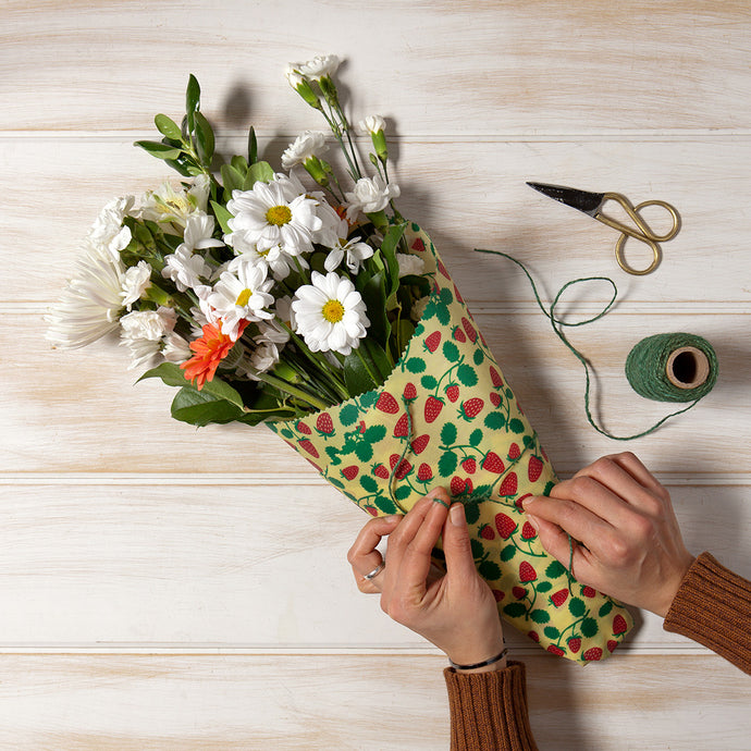 A yellow beeswax wrap with red raspberries and green stems printed, is wrapped around the bottom part of a flower bouquet and is used as an alternative to the plastic wrap they usually gift. Hands are shown wrapping with twine.
