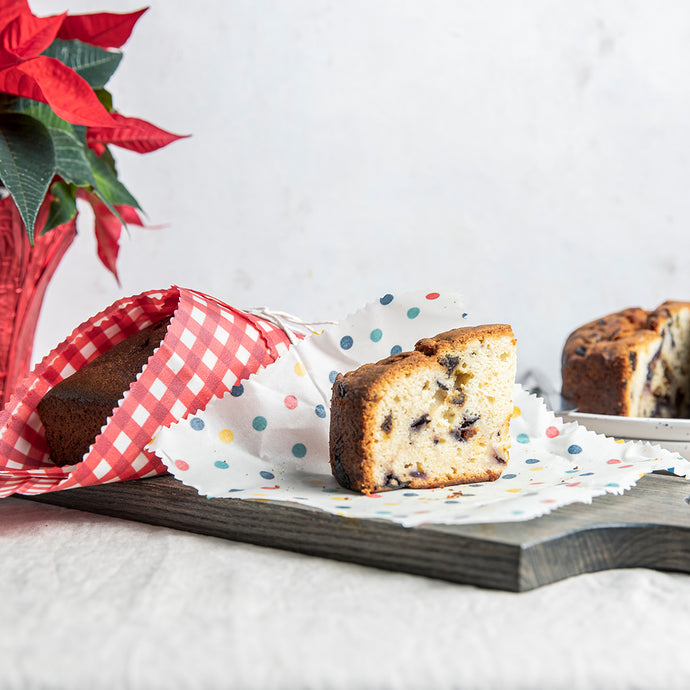 White Beeswax wrap with red and blue polkadots printed, wraps a piece of holiday fruit cake. Stationed on a grey cutting board with marble background. Behind is bread wrapped in a red gingham beeswax wrap.