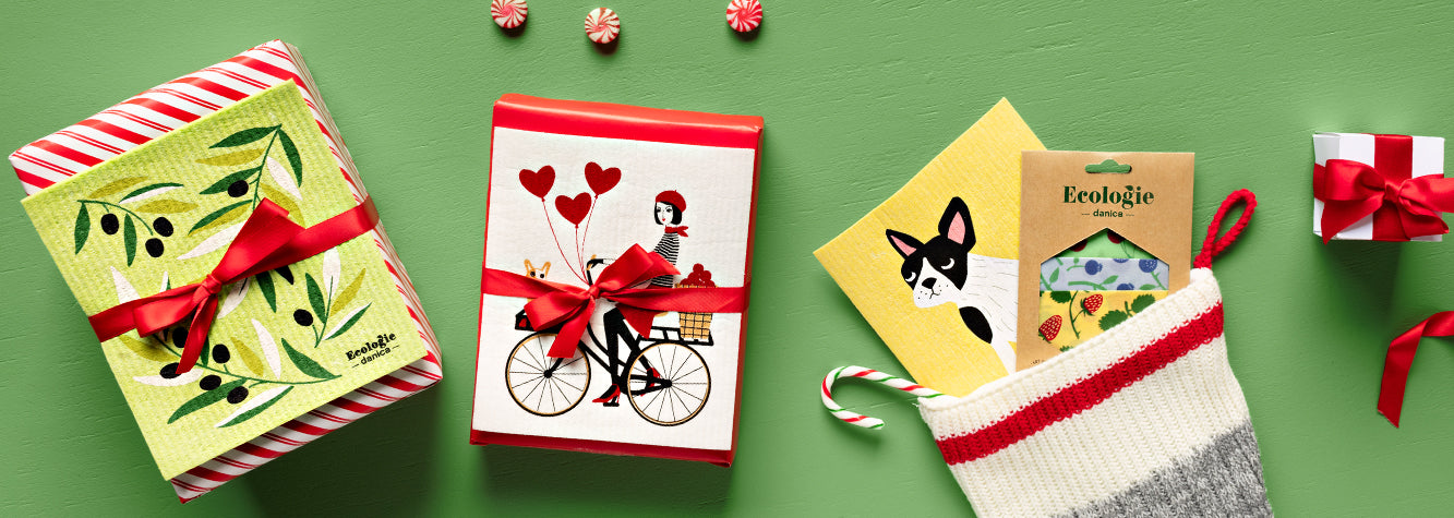 Green backdrop with a christmas stocking stuffed with a yellow sponge cloth with a French Bulldog printed on, and a set of beeswax wraps with berries & fruit printed. To the left is 2 wrapped gifts in red paper, with a sponge cloths tied on top.